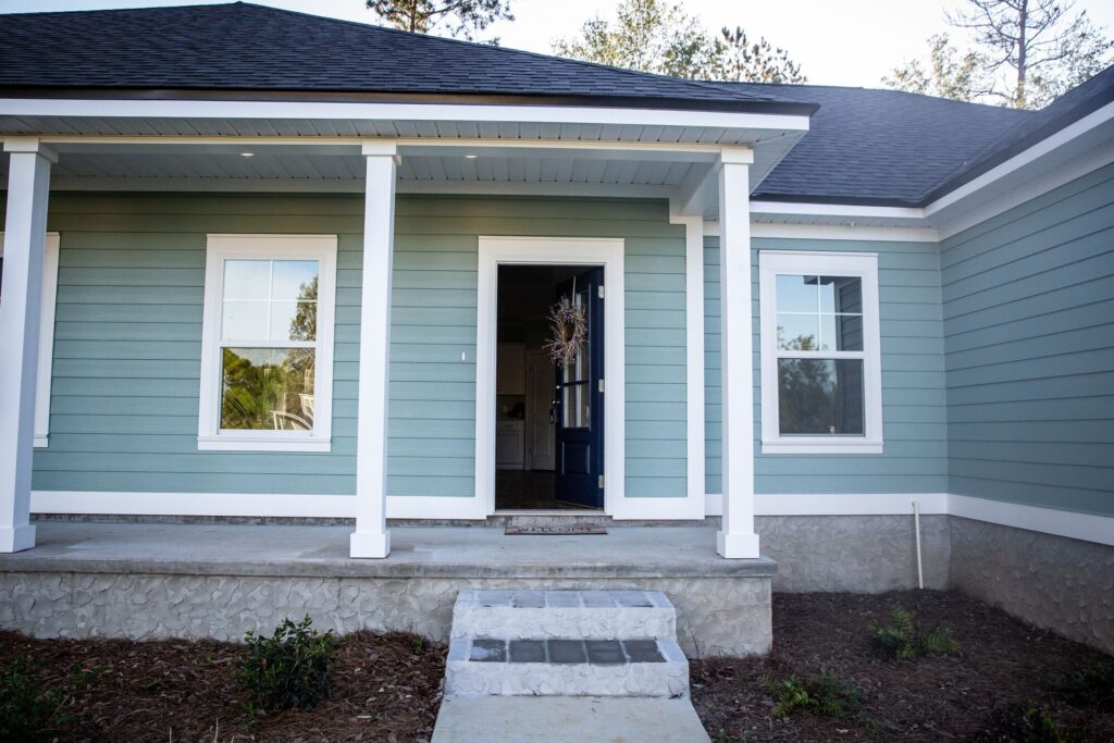 Front porch of a modern blue-green house featuring white trim, a welcoming dark front door, and decorative wreath, set against a sunny backdrop.
