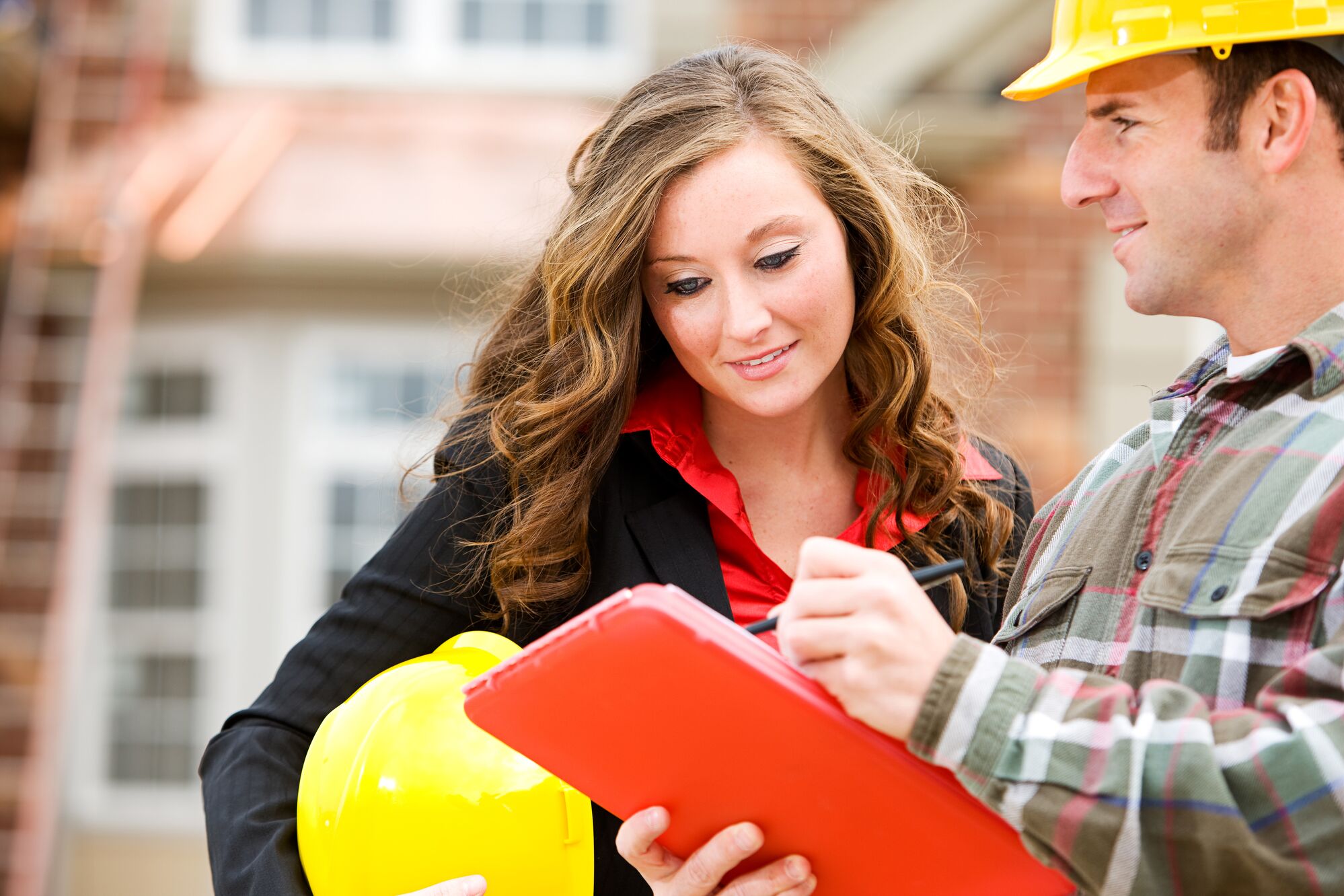 A woman in a black jacket and red blouse holds a yellow hard hat while reviewing notes with a man in a plaid shirt and hard hat. They discuss project details.