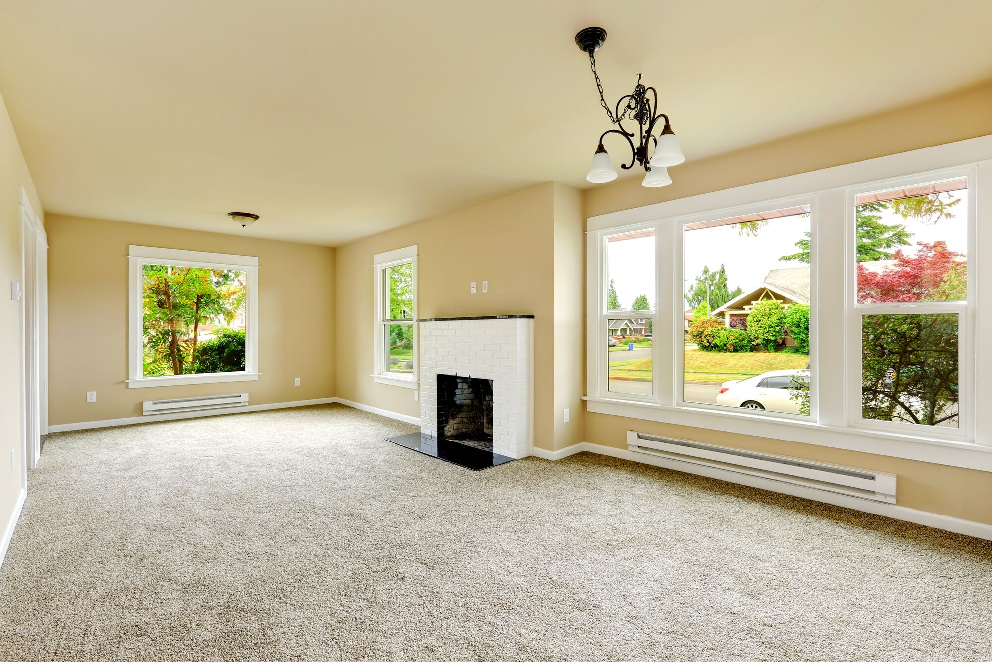 Bright, airy living room featuring large windows, a white fireplace, and beige walls. The spacious carpeted area invites relaxation.