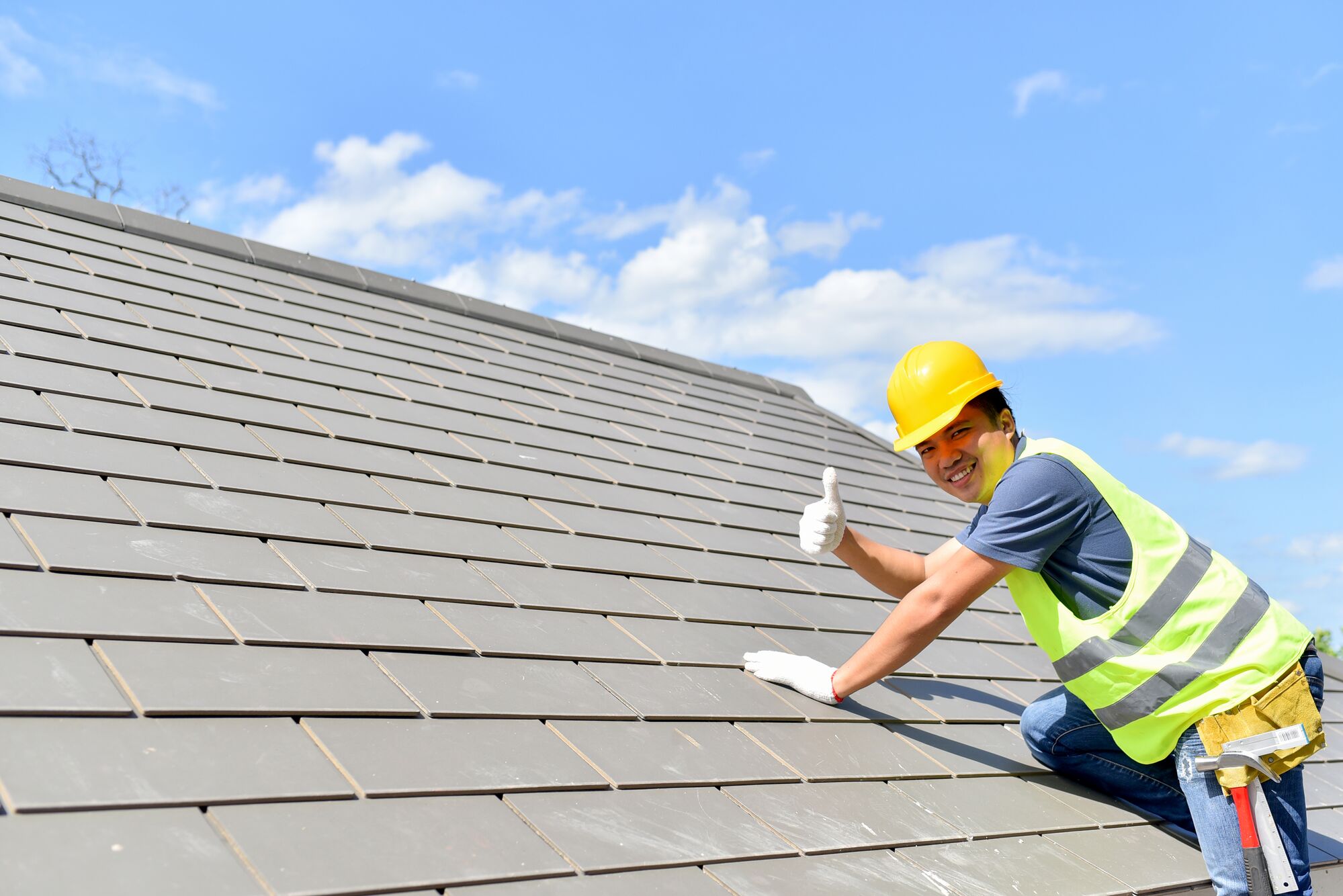 Roofer in a yellow hard hat and safety vest gives a thumbs up while working on a tiled roof under a clear blue sky.
