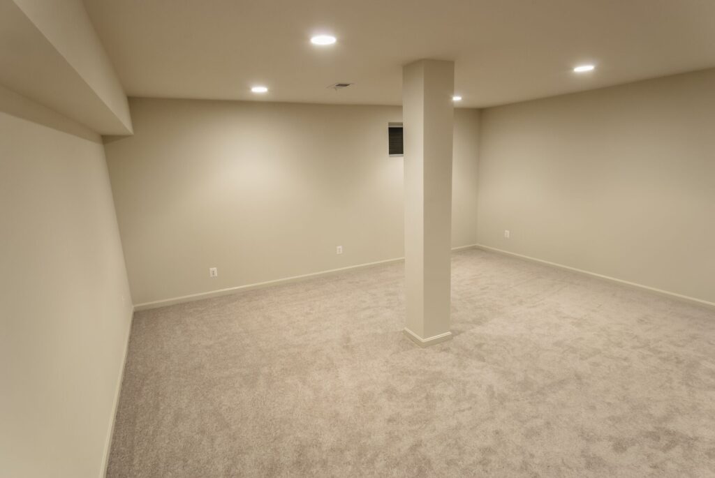 Spacious, empty basement room with beige carpet and neutral walls, featuring recessed lighting and a central support pillar. Ideal for flexible use.
