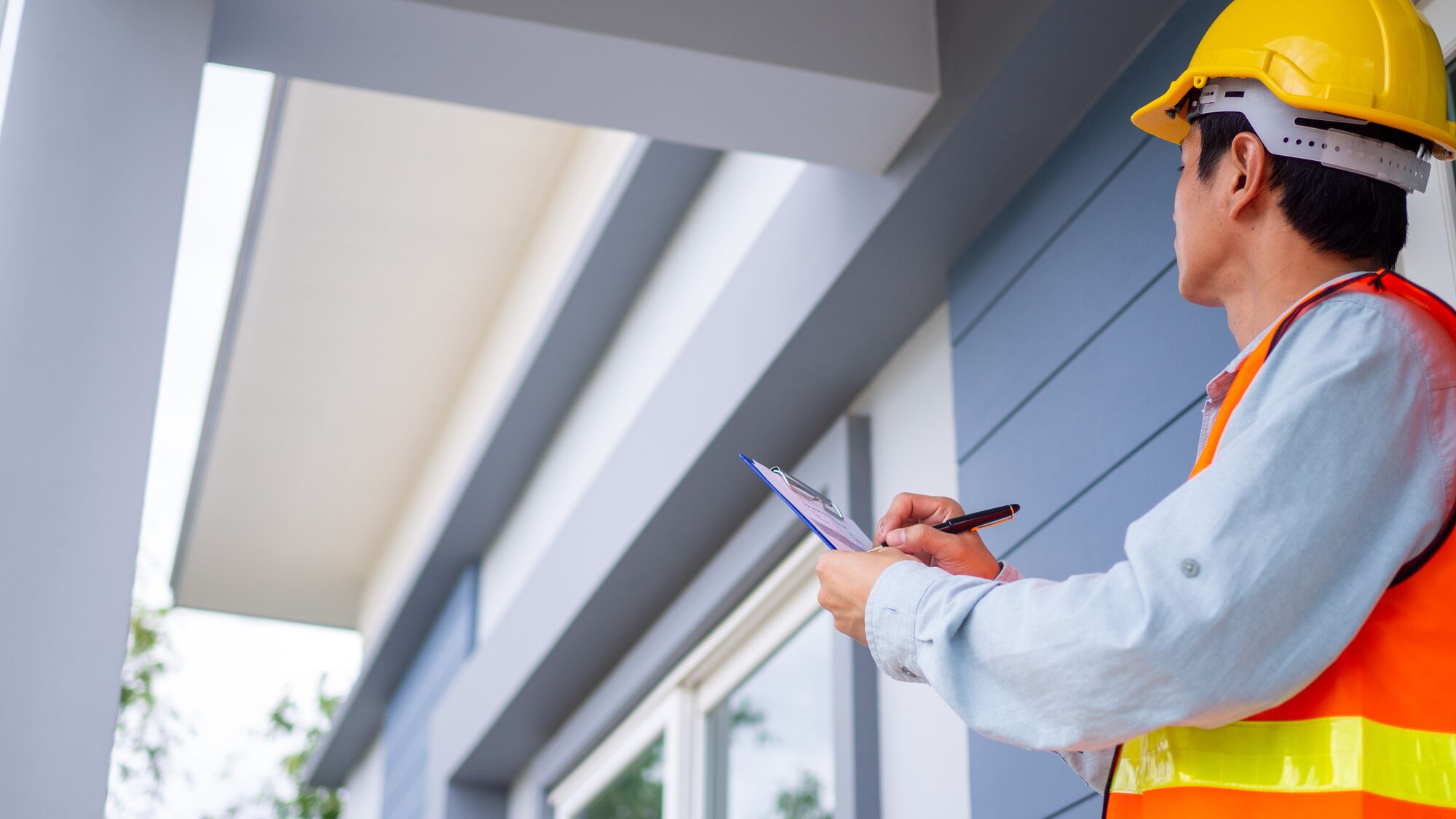 Construction worker in a hard hat and safety vest inspects a building, taking notes on a clipboard to ensure compliance and safety standards.