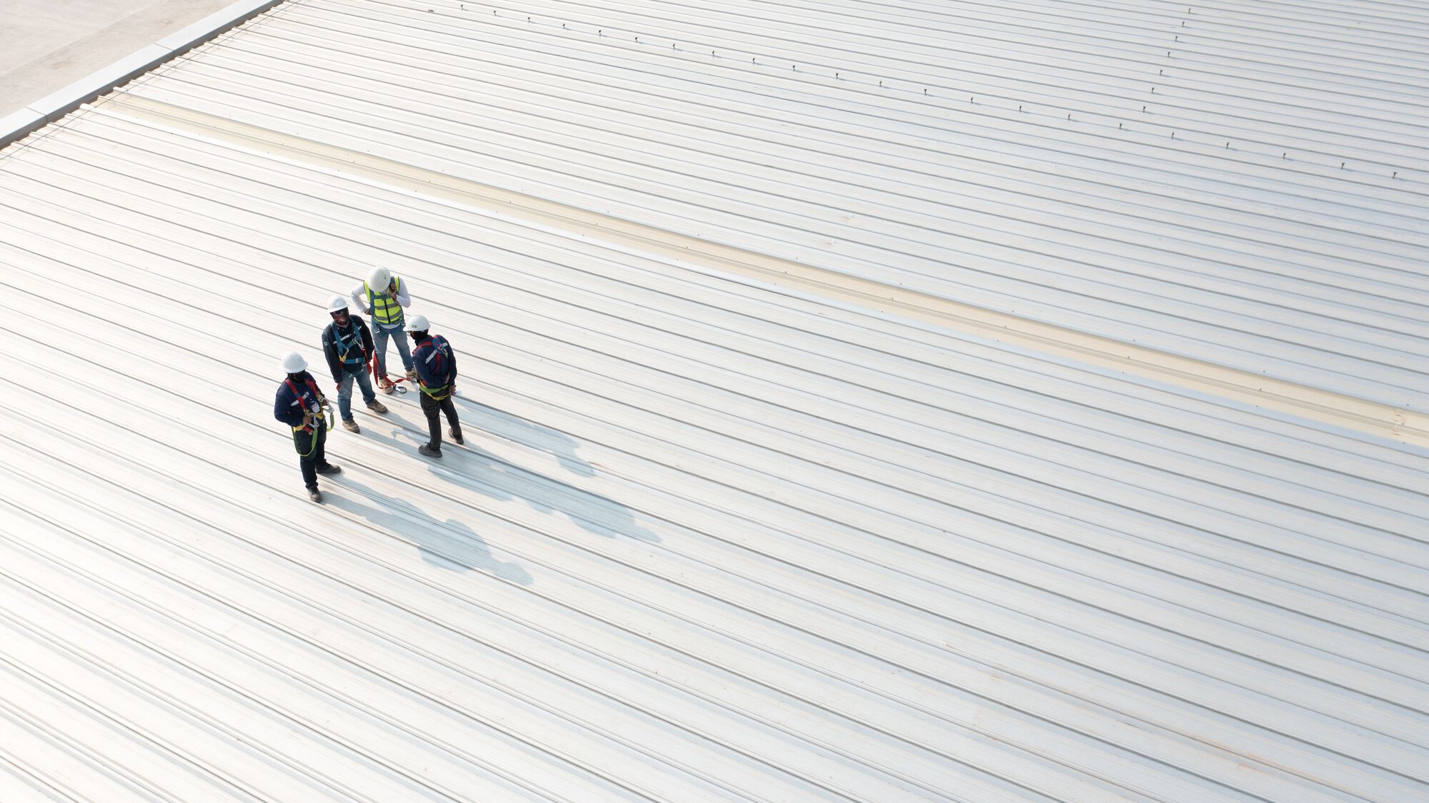 Four construction workers in helmets and safety vests discuss plans on a commercial roof, highlighting teamwork and safety in construction.