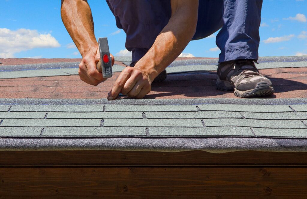 A worker is repairing a roof, using a hammer on shingles while kneeling. This image illustrates roofing maintenance and home repair.