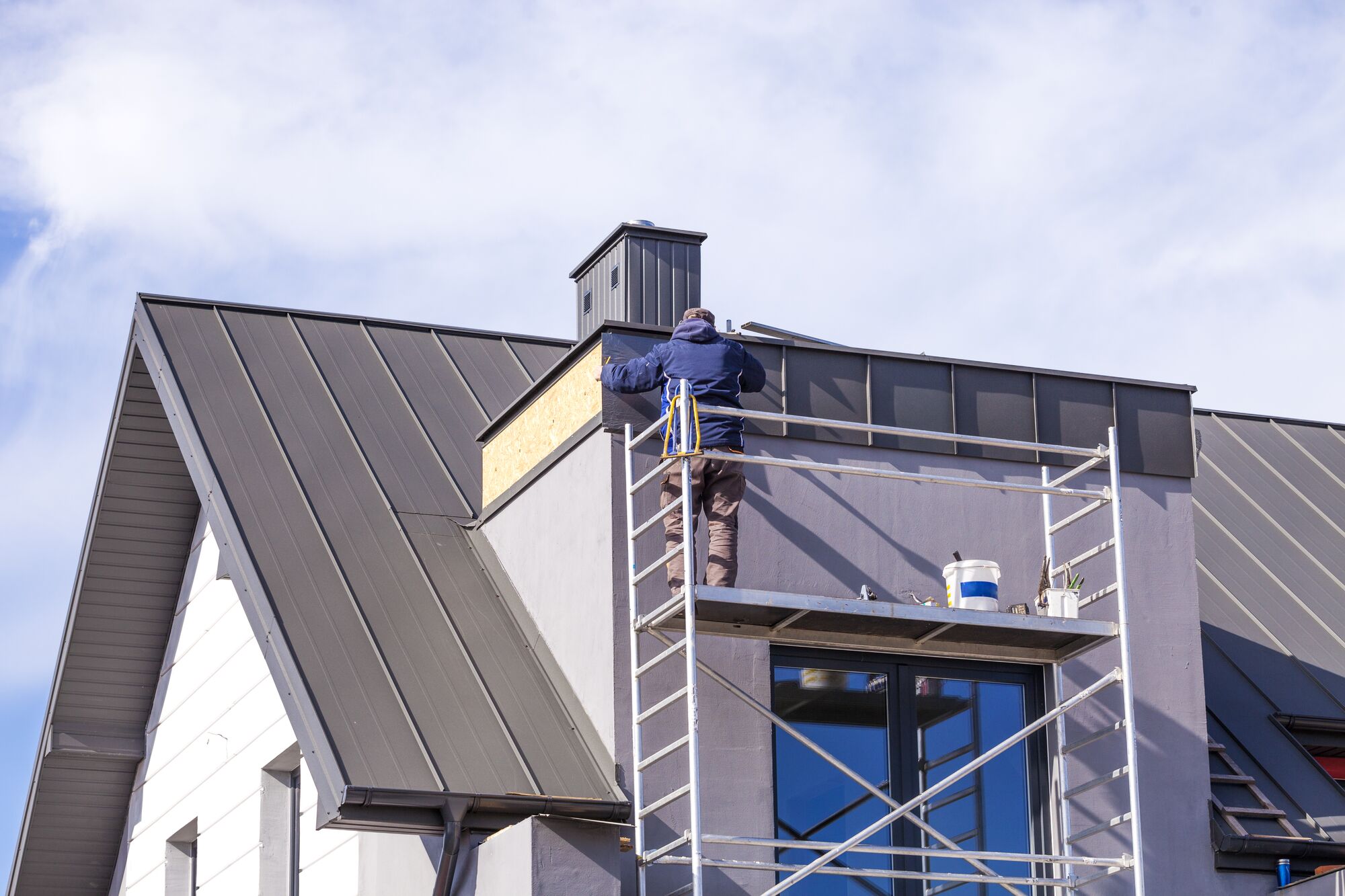 Worker on a scaffold making repairs to a metal-roofed building, showcasing home improvement and construction activities. Bright blue sky visible.