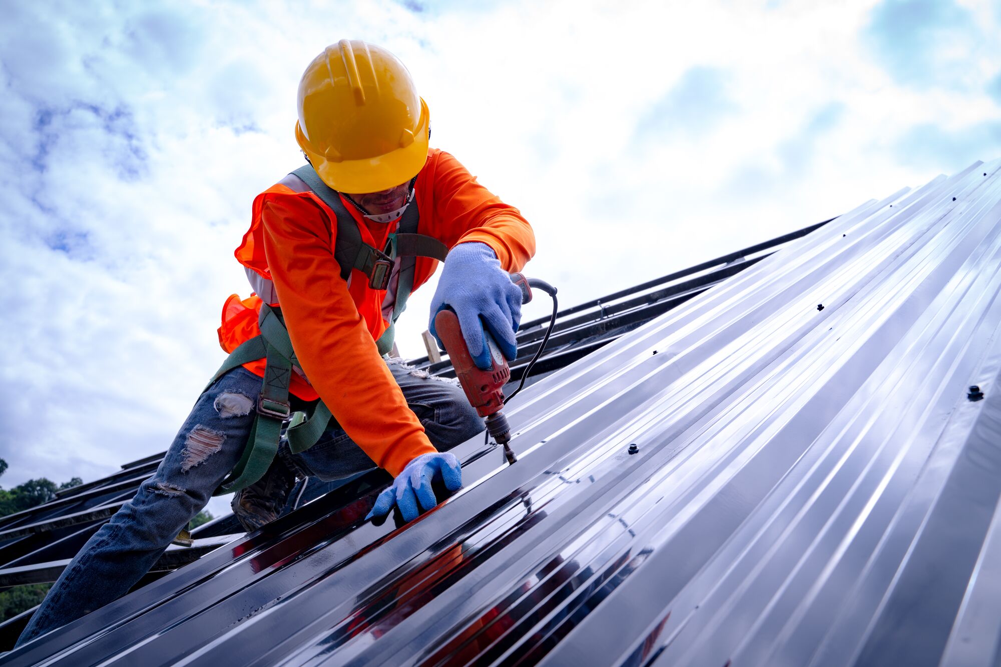 Construction worker in an orange shirt and hard hat drilling metal roofing panels while secured with a harness against a cloudy sky.