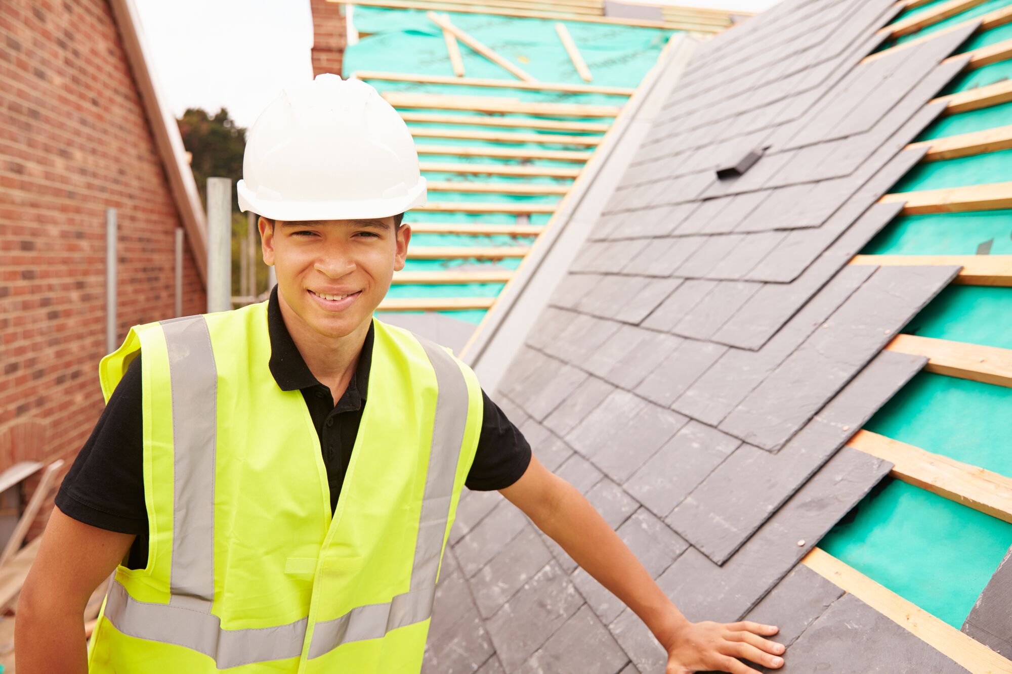Young construction worker in a hard hat and fluorescent vest smiles while standing on a sloped roof, showcasing slate shingles and construction materials.