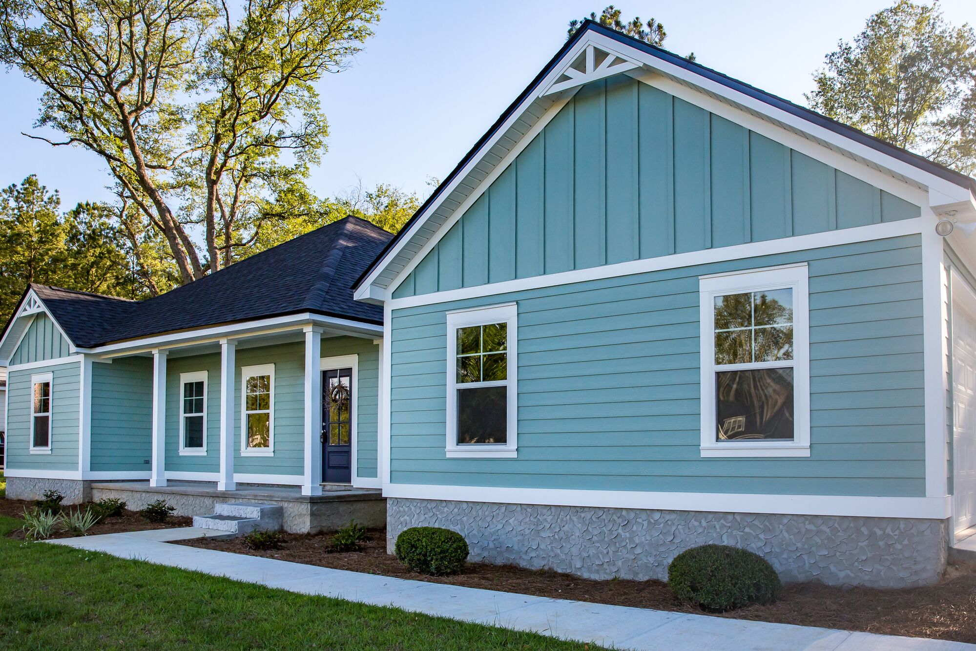 Modern light blue house with white trim, featuring a welcoming porch and landscaped front yard. Highlights a blend of contemporary design and nature.