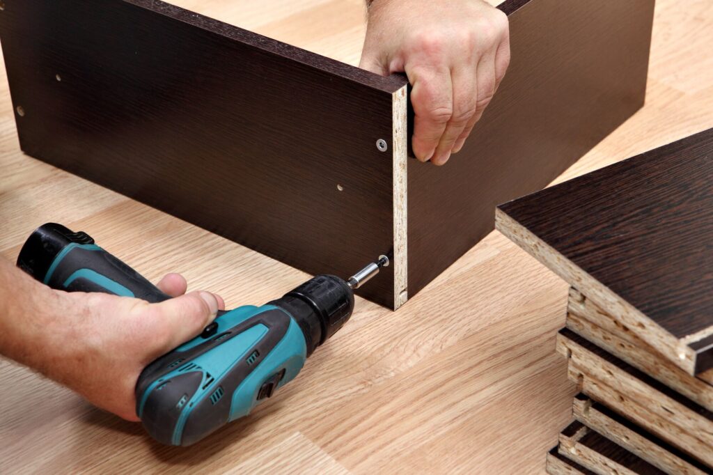 A hand using a power drill to assemble a dark wooden cabinet, with stacked pieces of the cabinet visible nearby. Relevant for DIY furniture assembly guides.