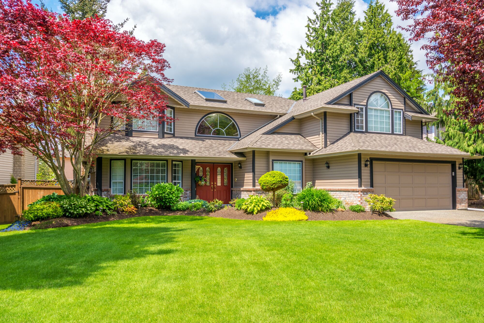 A two-story house with a welcoming façade features large windows and a red front door, surrounded by lush green lawn and colorful landscaping.