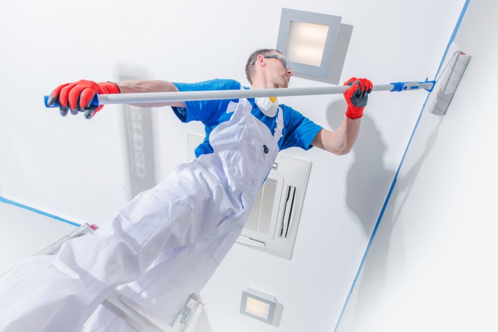 A painter in white overalls uses a long roller to paint the ceiling blue, focusing on detail while standing on a ladder.