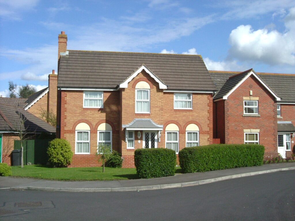 Two-story red-brick house with a pitched roof, surrounded by neatly trimmed hedges, located on a residential street under a clear blue sky.